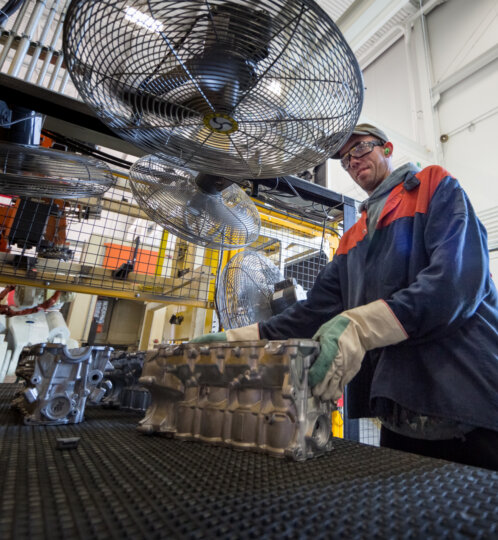 A worker in safety gear and gloves stands at a textured workbench, handling a metal engine component in a large industrial workshop with ceiling fans.