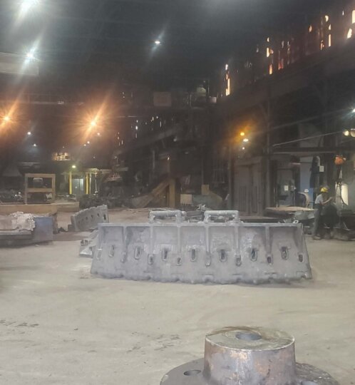 Nighttime industrial site with bright floodlights, gray concrete barriers, and large metal rings in the foreground; workers in hard hats visible in the background.