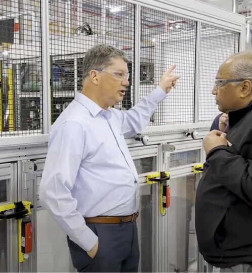 Two workers wearing safety glasses stand beside a mesh-fenced industrial machine in a bright factory; one gestures with a raised hand, the other listens.