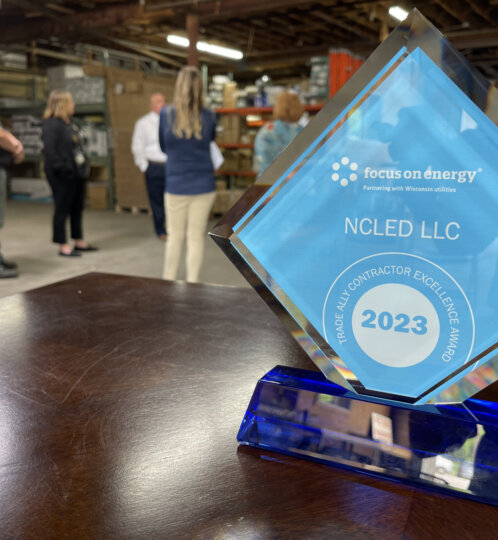 Blue diamond glass trophy on a wooden table, displaying Focus on Energy, NCLED LLC, 2023, and a circular award seal; group of people in a warehouse background.