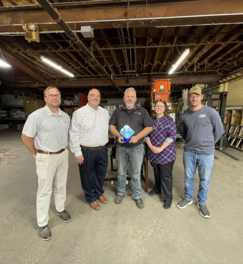 Five people stand in a warehouse; the center person holds a blue award, with shelves, ladders and industrial equipment in the background.