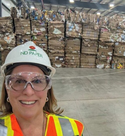 Person wearing a white ND PAPER hard hat, safety goggles, and a high-visibility vest, taking a selfie in a warehouse with tall stacks of cardboard bales.