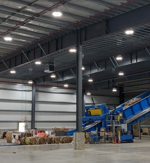 Industrial warehouse with a blue inclined conveyor sorting cardboard bales; stacks of bales and machinery under bright ceiling lights.