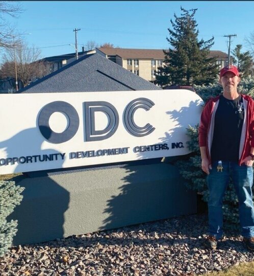 A person stands beside a large white sign reading 'ODC' and 'Opportunity Development Centers, Inc.' outdoors, wearing a red cap, red jacket, and blue jeans.