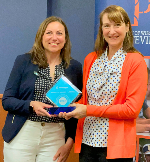 Two people indoors smiling as one in a navy blazer presents a blue glass 2023 award to the other who wears an orange cardigan over a polka-dot blouse.