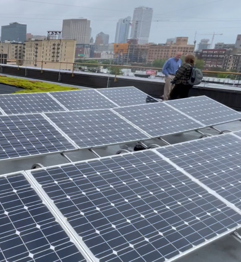 Rooftop covered with solar panels in rows; a railing edge with several people standing and walking, and a city skyline in the distance.