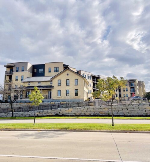 Modern beige apartment complex on a stone retaining wall, with trimmed trees in the foreground and a road in front beneath a cloudy sky.