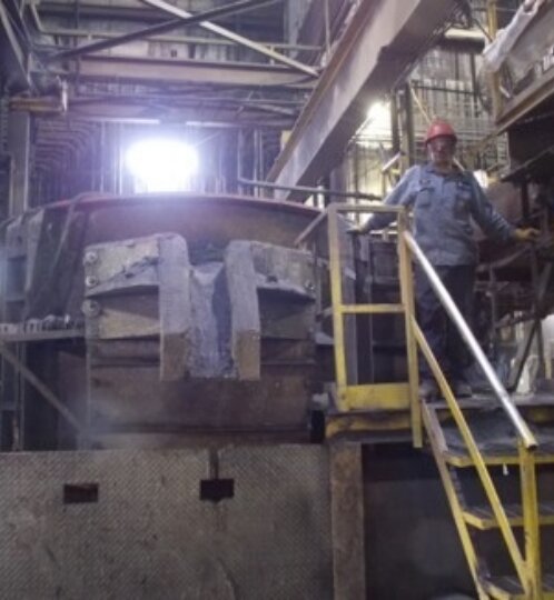 A person in a red hard hat stands on a yellow-railed stair platform beside a large dusty industrial machine in a factory.