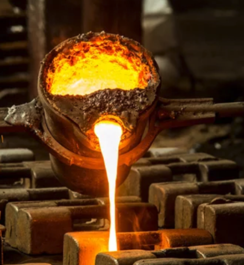 Molten metal pours from a crucible into a mold in a metal foundry, casting a bright orange glow as sparks fly and heavy equipment surrounds the scene.