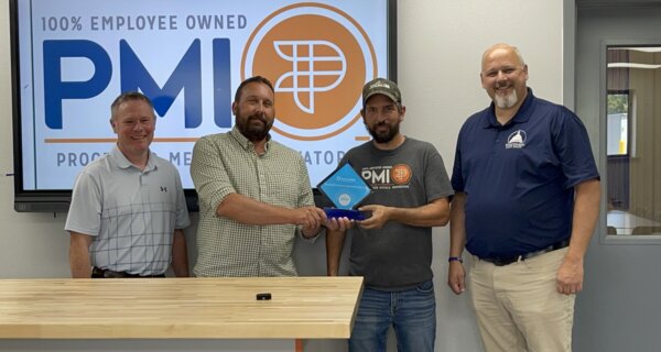 Four adults stand in an office lobby; two center figures shake hands while presenting a blue trophy, with a large PMI logo on a screen behind them.