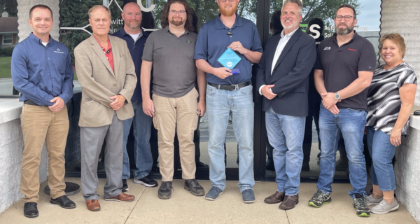 Nine adults stand in a row outside a storefront, smiling; the center person holds a blue award plaque.