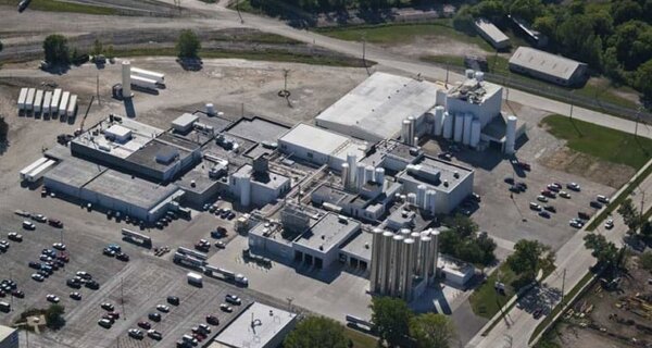 Aerial view of a large industrial facility with white silos, flat-roofed buildings, loading areas, and a spacious parking lot.