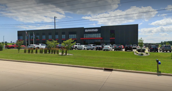 A modern dark-gray office building with red trim sits behind a long parking lot full of cars, with green lawns, small trees, and flagpoles under a blue, partly cloudy sky.