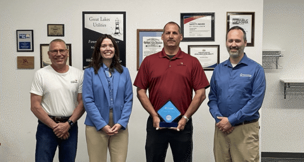 Four people pose indoors in front of a wall of awards; the person in a red polo holds a blue award plaque, flanked by others in casual/business attire.
