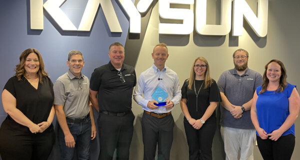 Seven people stand in an office lobby under a large KAYSUN sign; the center person holds a blue award while others smile.