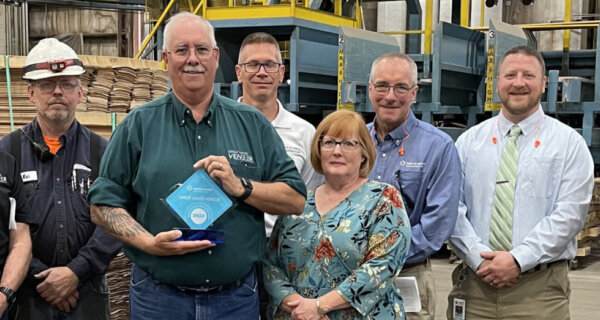 Group of professionals posing in a veneer warehouse; center person holds a blue award plaque, with stacks of plywood and shelving in the background.