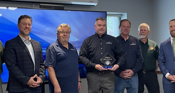Eight people in business casual attire pose in a conference room; the center person holds a glass trophy labeled 2025, with a blue-screen backdrop.