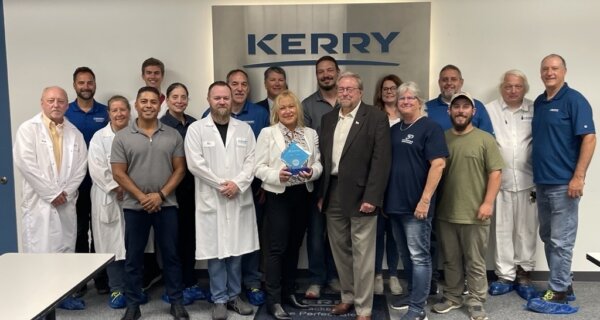 Group of staff posing in a Kerry facility in front of a large Kerry sign; several wear white lab coats and blue shirts, one person holds a blue award plaque.