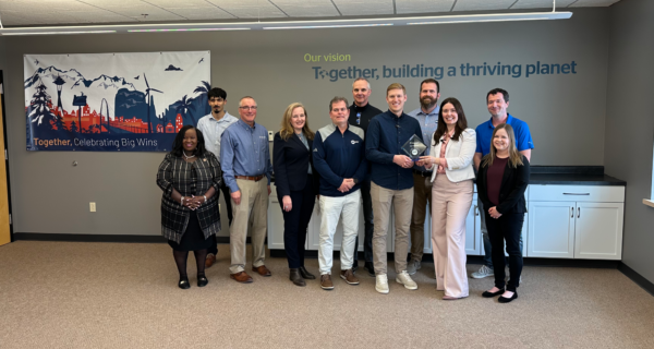 Diversified group of colleagues pose in an office; left banner features skyline and wind turbines, and two people hold a glass award.