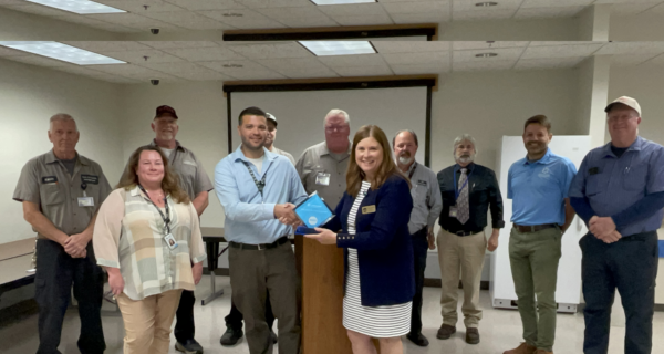 Group of ten in a conference room; two center people shake hands as a blue award plaque is exchanged, while others smile near a podium.