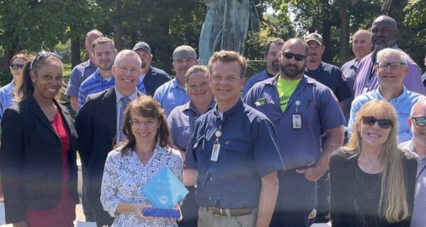 A diverse group of professionals outdoors in a park, smiling for a group photo; a front-center person holds a blue glass trophy with a statue in the background.