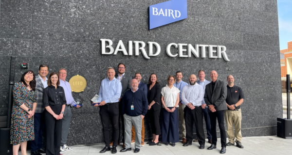 Group of professionals posing outside the Baird Center, holding a LEED Gold plaque and a trophy, in front of a dark granite wall.