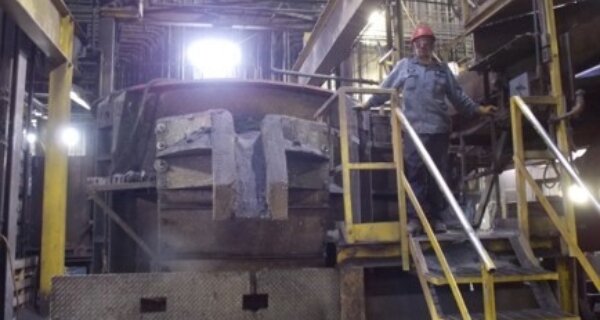 A person in a red hard hat stands on a yellow-railed stair platform beside a large dusty industrial machine in a factory.