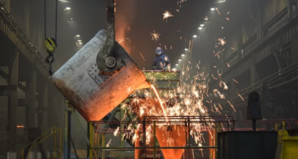 Foundry interior with a crane bucket pouring molten metal, sparks flying as a worker in protective gear stands on a platform above.