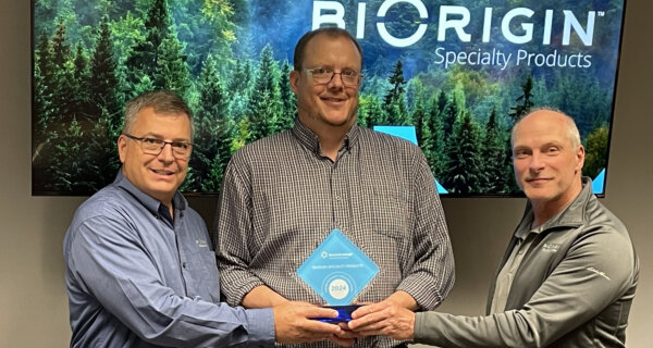 Three people indoors, smiling, jointly holding a blue diamond-shaped award marked 2024, with a forest-themed screen and company branding in the background.