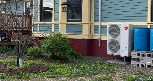 Teal house with yellow trim, wooden deck left, AC unit, blue rain barrels on blocks, and a small garden with a tree and red flowers.