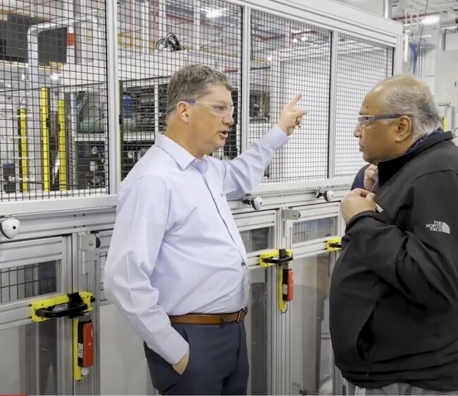 Two workers wearing safety glasses stand beside a mesh-fenced industrial machine in a bright factory; one gestures with a raised hand, the other listens.