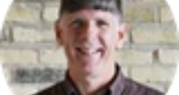 Portrait of a smiling person with short dark hair wearing a brown shirt, posed in front of a brick wall.