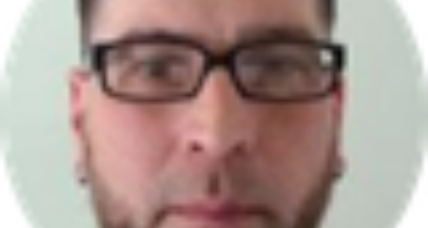 Circular portrait of a person with short dark hair and light stubble, wearing rectangular glasses, looking at the camera against teal background.