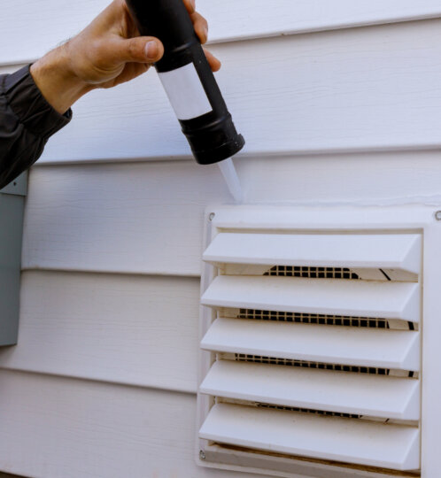 Hand holds a spray nozzle and sprays water at a white exterior vent on the side of a house with horizontal siding.