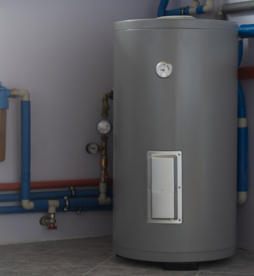 Industrial utility room featuring a tall gray water heater, blue and red pipes, gauges, and a brown filter housing on the left.