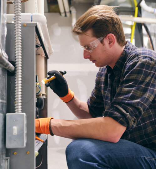 Person wearing safety goggles and orange gloves kneels beside electrical equipment, using a tool on a panel in a workshop or lab.