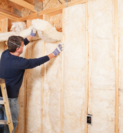 Construction worker wearing a mask and gloves installs fiberglass insulation between wooden studs, standing on a ladder in a partially finished wall.