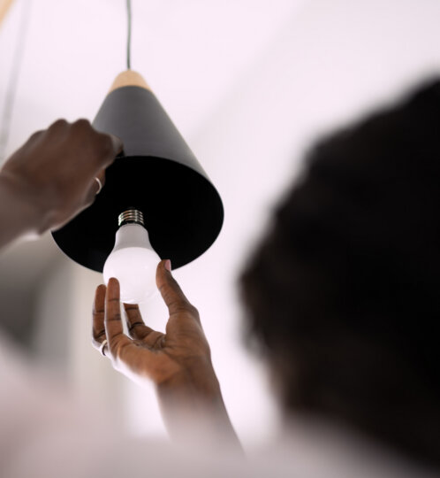 Close-up of hands installing a light bulb under a black cone-shaped pendant lamp, with a blurred figure in the foreground.