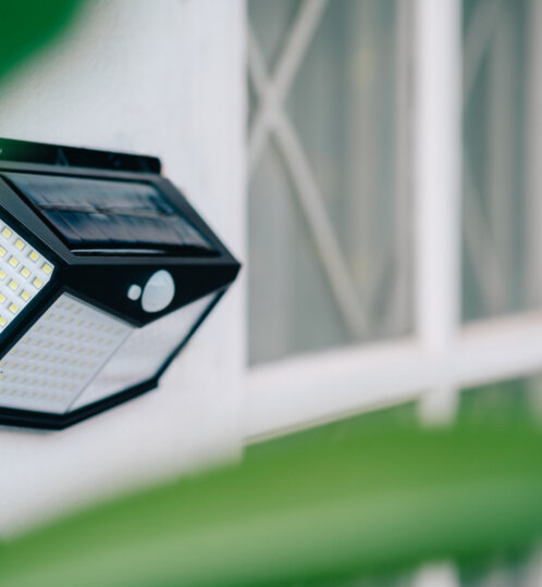 Outdoor black geometric LED wall light with a solar panel on top, mounted on a white wall near a window, with green leaves in the foreground.