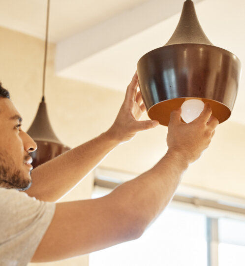 Person with short dark hair in a light gray shirt reaches up to install a brown pendant light, inserting a bulb beneath its orange interior shade.