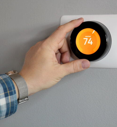 Close-up of a hand adjusting a circular smart thermostat on a white wall plate; orange display reads 74 and HEATING, with a blue plaid sleeve and silver mesh watch.