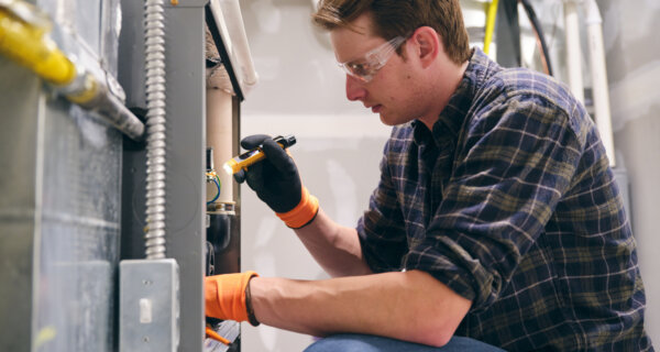 Person wearing safety goggles and orange gloves kneels beside electrical equipment, using a tool on a panel in a workshop or lab.