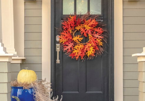 Front porch with a dark door adorned by a bright autumn wreath of red, orange, and yellow leaves; a yellow pumpkin rests on a blue pot amid dried grasses.
