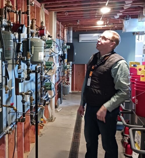 Person wearing a vest and lanyard stands in a utility room filled with exposed pipes, gauges, and valves along a wall, looking up at the ceiling.