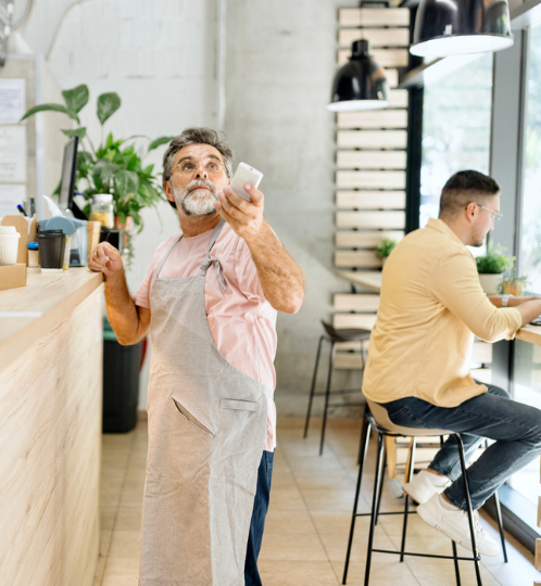 A cafe scene: older person in a pink shirt and light apron holds a smartphone, gesturing, while another person in yellow sits at a window seat.