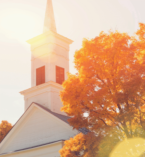 White church with a tall steeple beside a large orange autumn tree, bathed in warm sunlight from the left.