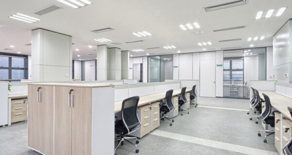 Bright, modern open-plan office with long rows of light-wood desks, black ergonomic chairs, white walls, glass doors, and bright overhead lighting.