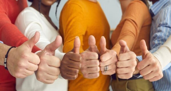 A diverse group in colorful outfits gives thumbs up toward the camera, signaling teamwork and positivity.