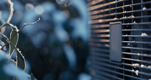 Close-up of frosty leaves along a branch beside an outdoor AC unit grille, with cool blue tones and a blurred background.