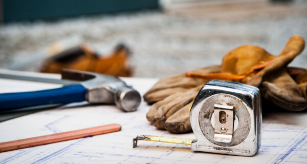 Close-up of construction tools on blueprints: tape measure, gloves, hammer, pencil, and ruler on a drafting table.